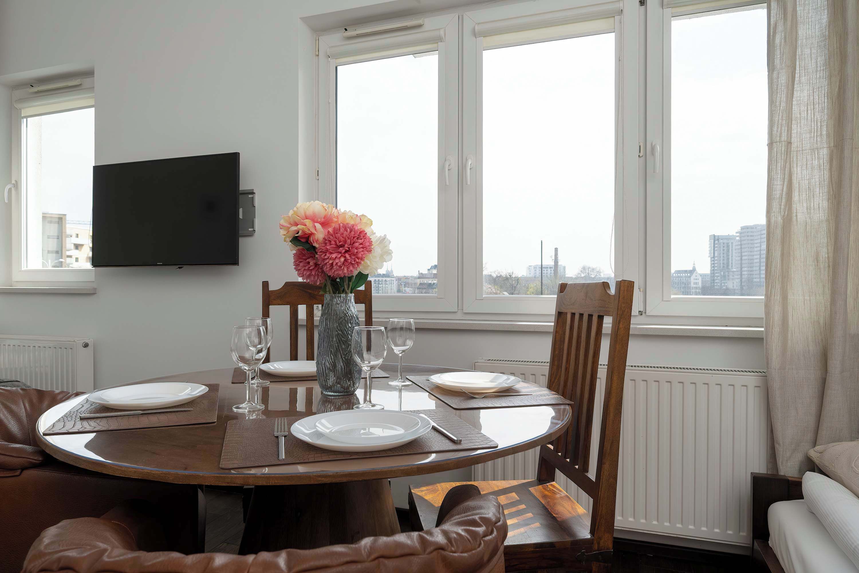 Ein Essbereich mit rundem Tisch, Stühlen, einer Vase mit Blumen und Blick auf die Stadt durch große Fenster.