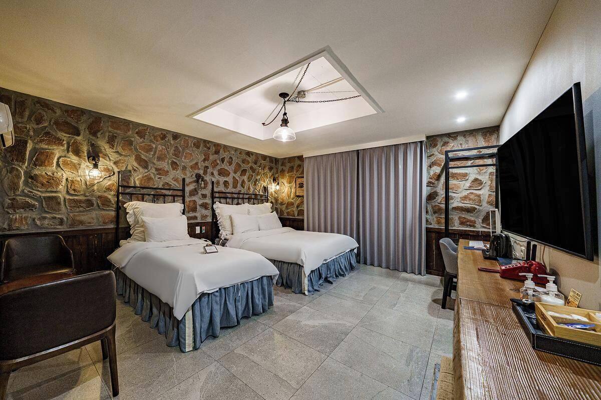 Camera d'albergo con due letti, una scrivania, una sedia, una televisione e un muro in pietra.
