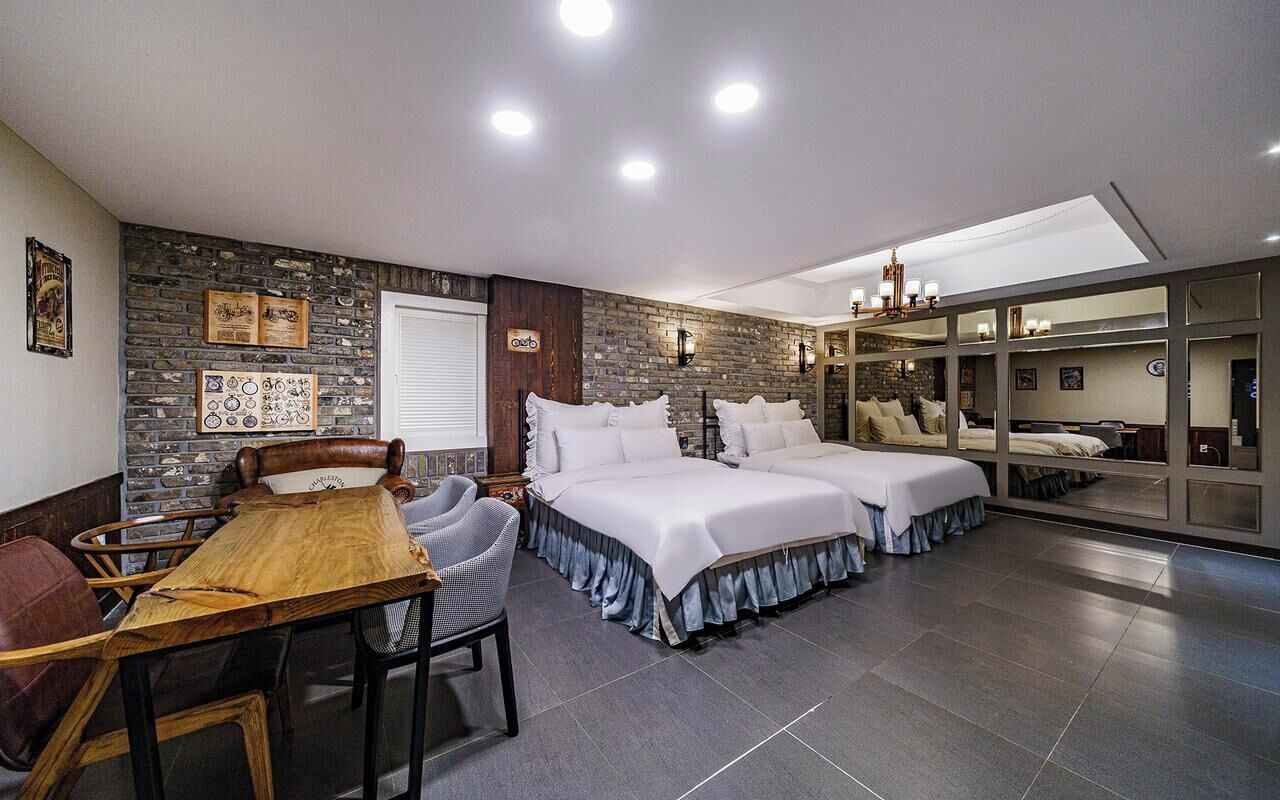 Una habitación de hotel moderna con pared de piedra, dos camas, un área de comedor con una mesa y sillas de madera, y un espejo grande.