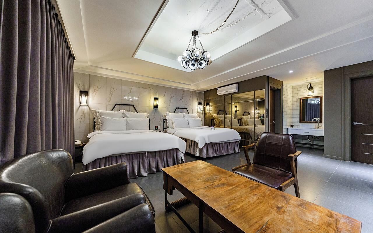 A hotel room with two beds, a wooden table, leather chairs, a chandelier, and a bathroom with a mirror and sink.