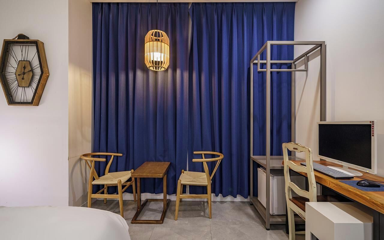 A room with a bed, a desk with a computer, two chairs, a small table, a wall clock, and a curtain.