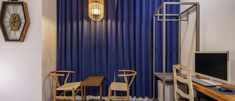 A room with a bed, a desk with a computer, two chairs, a small table, a wall clock, and a curtain.