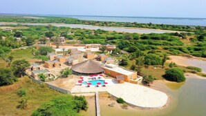 Vista aérea de un resort con varias construcciones, una piscina con tumbonas y una masa de agua al fondo.