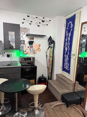 Une cuisine compacte équipée d’un four à micro-ondes, d’un grille-pain et d’une machine à café. On y trouve une petite table à manger avec des tabourets. Un aspirateur est placé près de la porte.