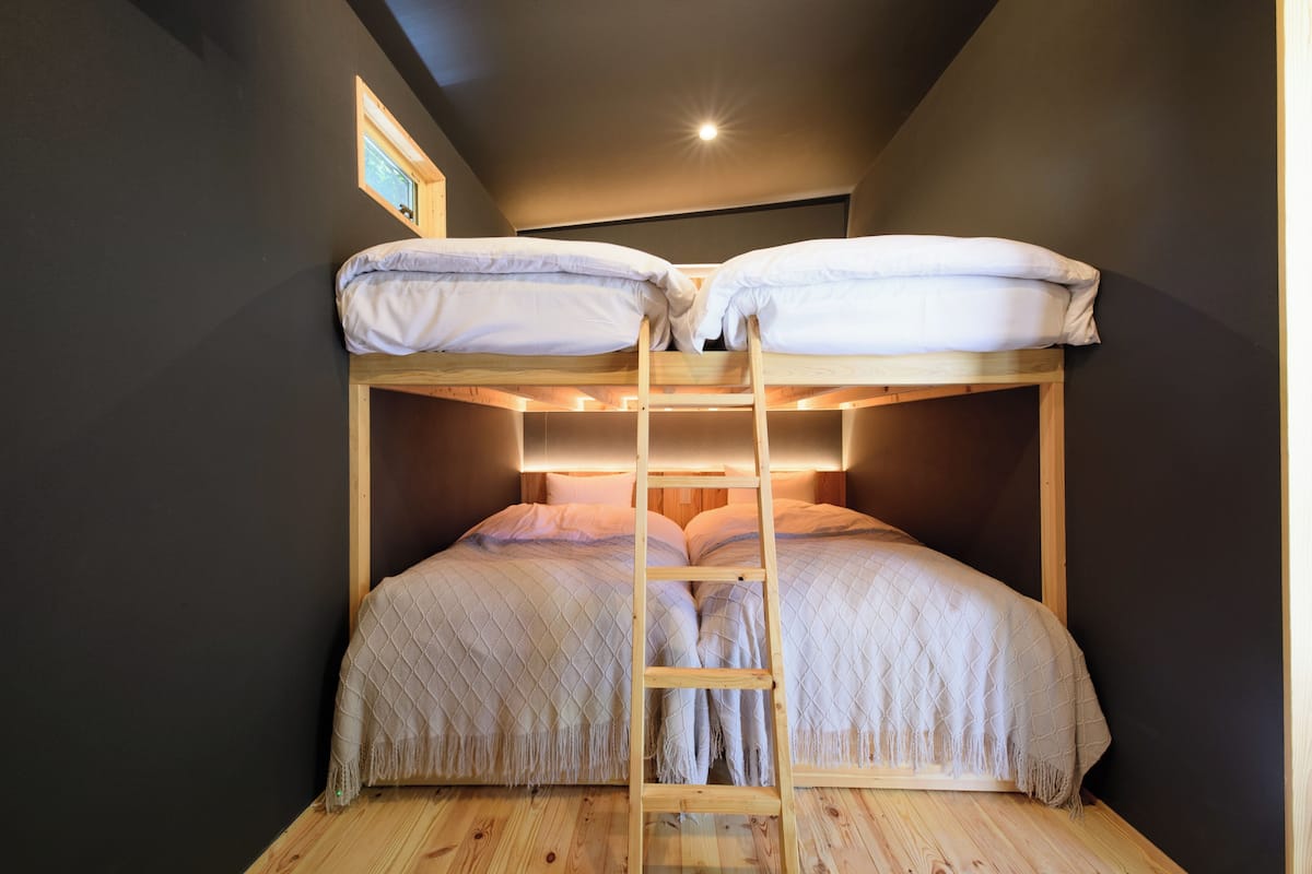 Un lit superposé avec une échelle en bois, une petite fenêtre et un luminaire fixé au mur.