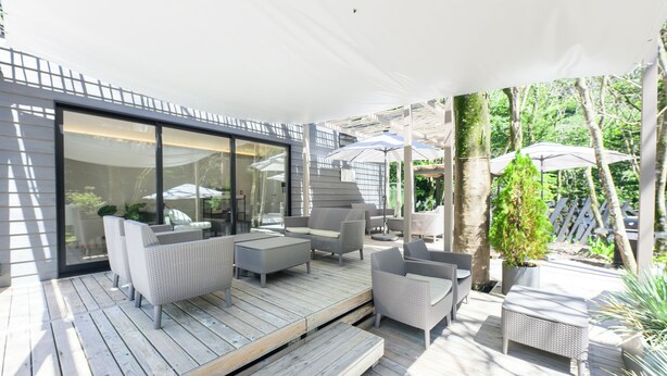 Een overdekte terras met rieten meubilair, een houten terras en een glazen deur die uitkomt op een tuin.