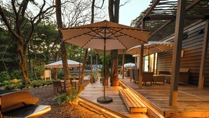 Een terras buiten met houten banken, parasols en een barbecueplaats.