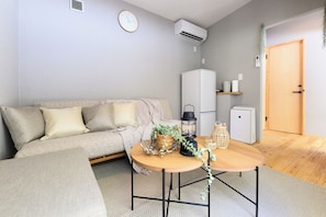 A living room with a sofa, a round wooden table, a small cabinet, a clock, and an air conditioning unit. - THE GUEST VILLAGE Hakone Ashinoko (Hakone)
