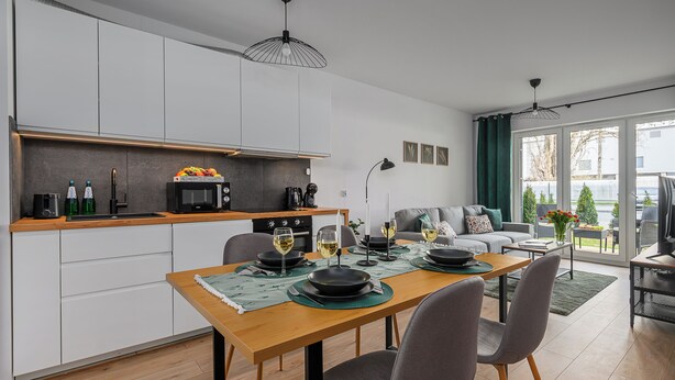 A modern kitchen with white cabinets, a dining table set for two, and a view of the outdoors through large windows. - Lumina apartment with patio and parking (Łódź)