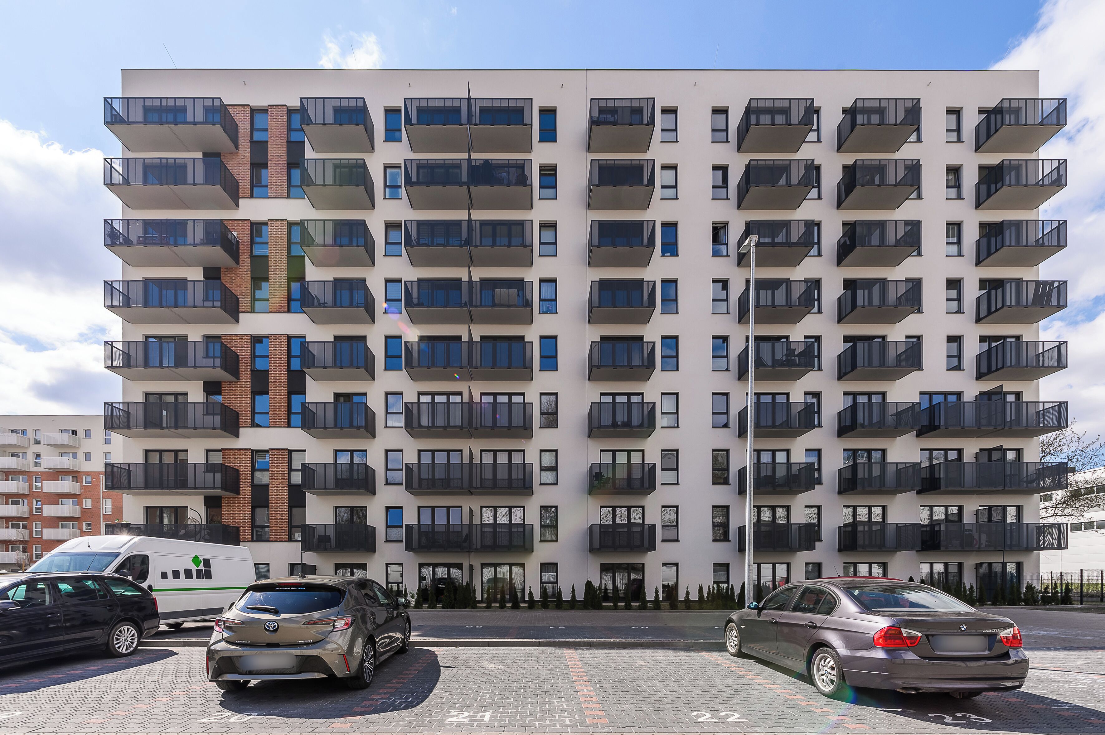 A multi-story residential building with balconies, a parking area, and a white van.