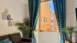 A living room with a sofa, a window with curtains, a bookshelf, and a potted plant. - Casa di Ada (Imperia)