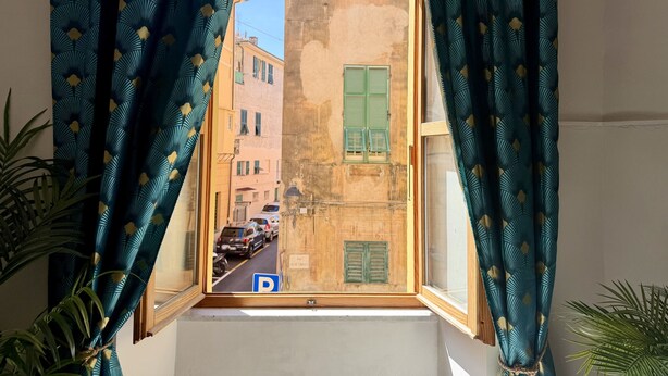 A window with patterned curtains, a view of a street with parked cars, and buildings with green shutters. - Casa di Ada (Imperia)