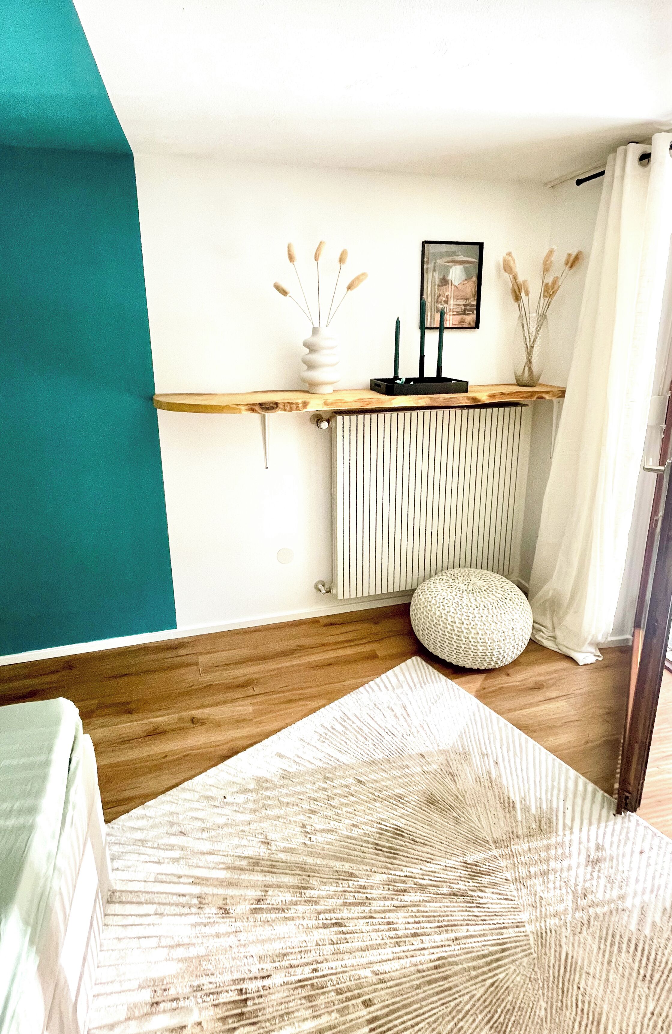 A room with a wooden floor, a white radiator, a teal accent wall, a small round pouf, a white vase with dried plants, and a framed picture on the wall.