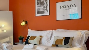 Ett hotellrum med en säng, vita kuddar, en inramad bild av en bil, en inramad Prada-logotyp och en sänglampa.