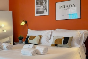 A hotel room with a bed, white pillows, a framed picture of a car, a framed Prada logo, and a bedside lamp. - Relax, Balcony & Pool Retreat (Numana)