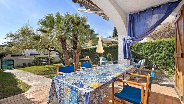 A patio with a table covered in a patterned tablecloth, wooden chairs, a palm tree, and a pergola with blue curtains. - Casita Abrilcon with Garden (Cap Roig)
