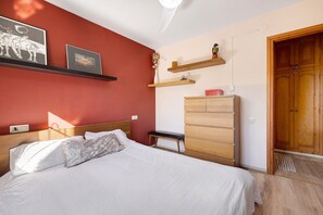A bedroom with a bed, a wooden dresser, a bench, and wall-mounted shelves with pictures and a small plant. - Casita Abrilcon with Garden (Cap Roig)