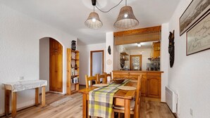 A dining area with a wooden table, chairs, and a checkered tablecloth. There is a kitchen in the background with wooden cabinets and a hanging light fixture. - Casita Abrilcon with Garden (Cap Roig)