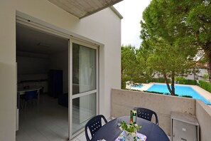 A balcony with a table, chairs, a glass door leading to a kitchen, and a swimming pool in the background. - Two level flat with balcony (Lignano Sabbiadoro)