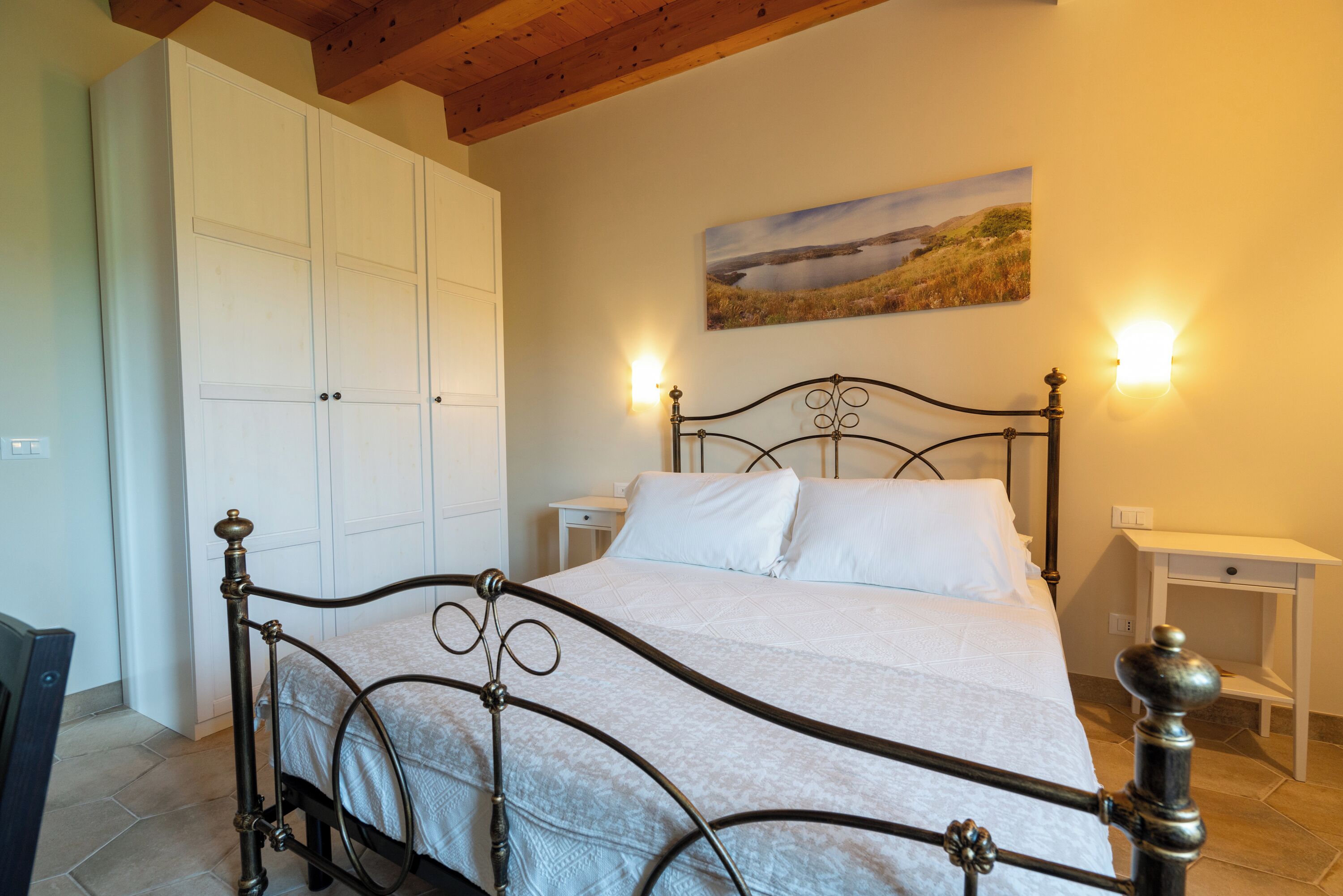 Een slaapkamer met een bed, een witte kledingkast, nachtkastjes en een landschap op de muur.