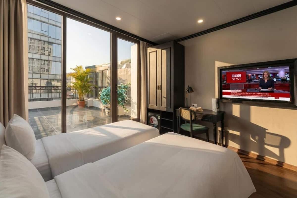 A hotel room with two beds, a TV displaying the BBC News channel, a desk with a chair, and a view of the outside balcony.