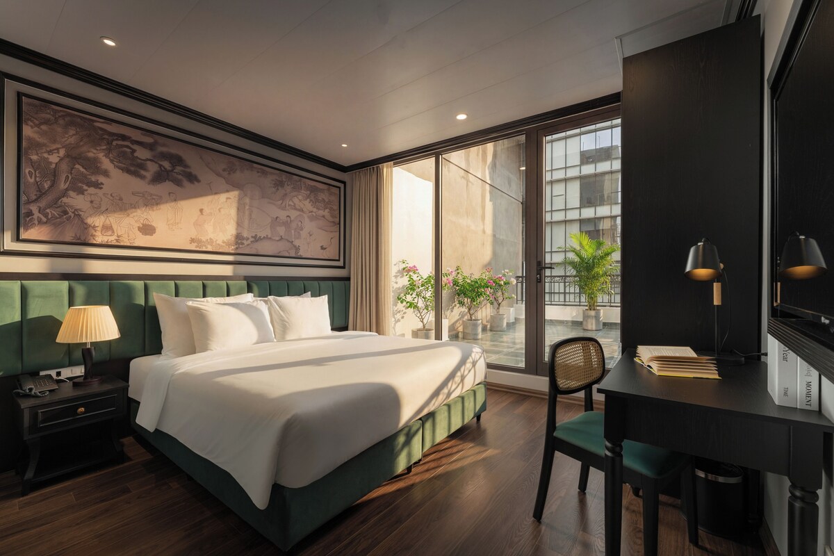 A modern hotel room with a large bed, a desk with a chair, a large window, and a painting on the wall.
