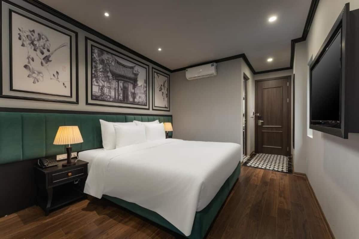 A hotel room with a large bed, a nightstand with a lamp, framed artwork on the wall, an air conditioning unit, a wooden door, and a television mounted on the wall.