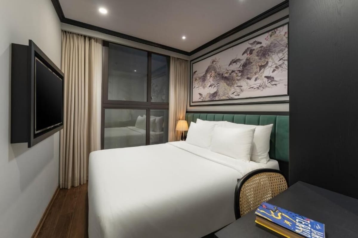 A hotel room with a bed, a television, a window with curtains, a wall-mounted artwork, and a table with books.