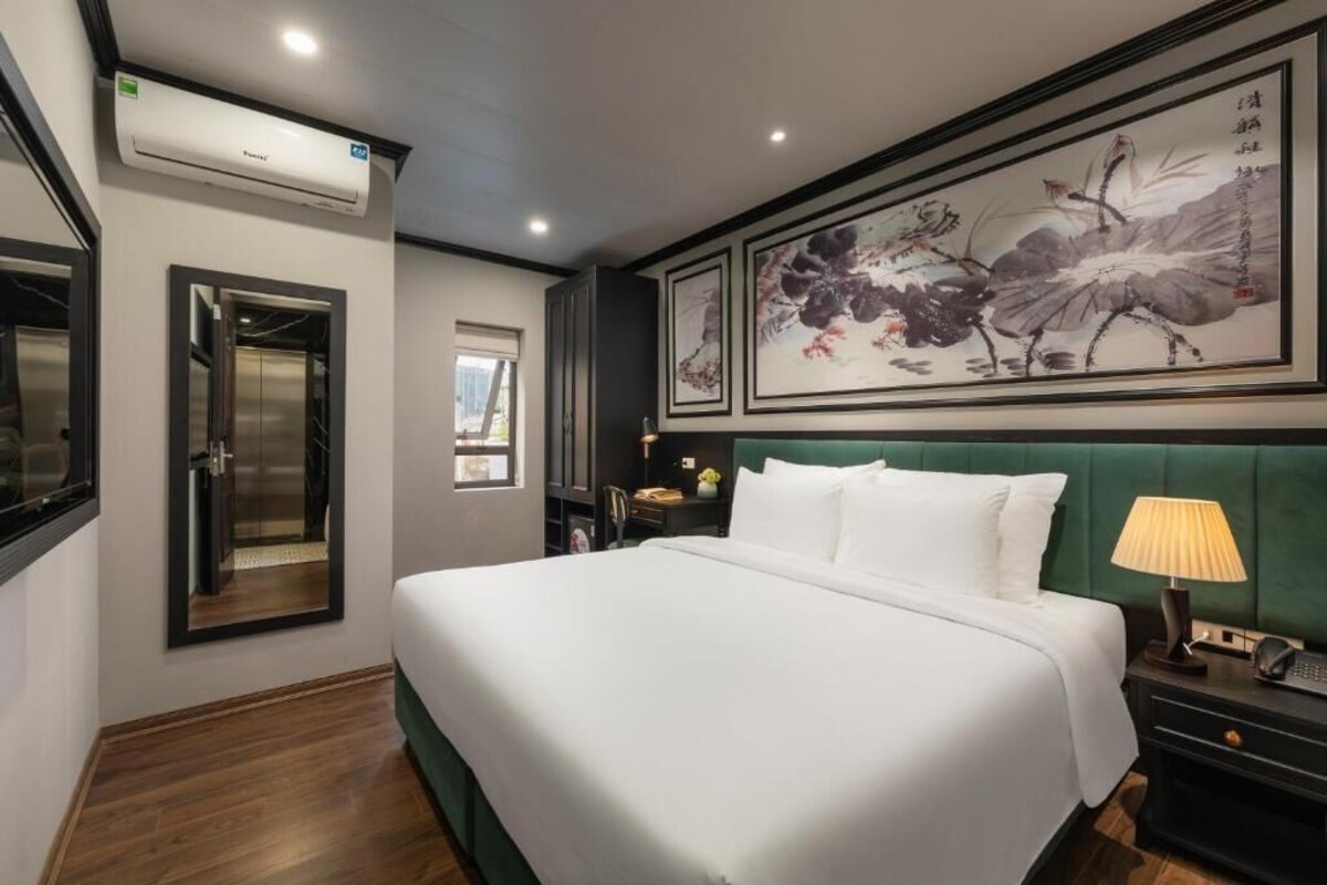 A hotel room with a bed, a desk, a chair, an air conditioning unit, and artwork on the wall.