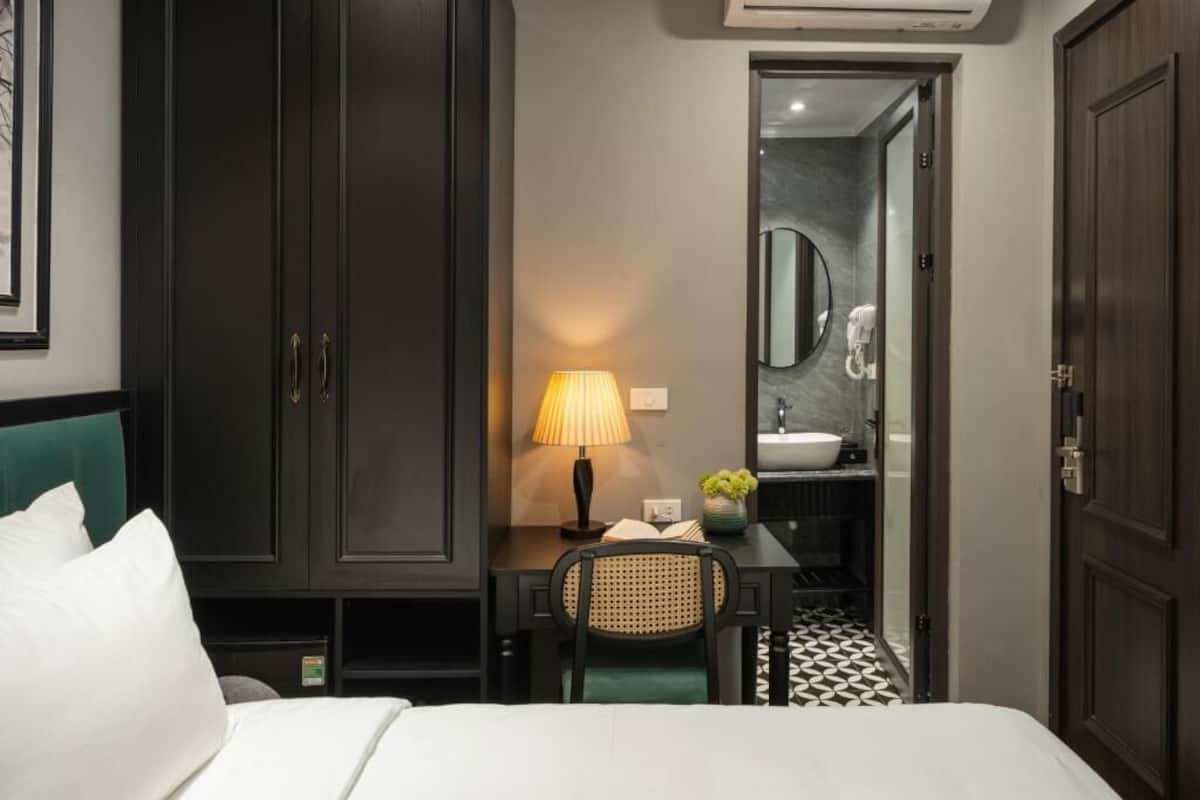A hotel room with a bed, a desk with a lamp, a chair, a small table with a vase, and a bathroom visible through an open door.