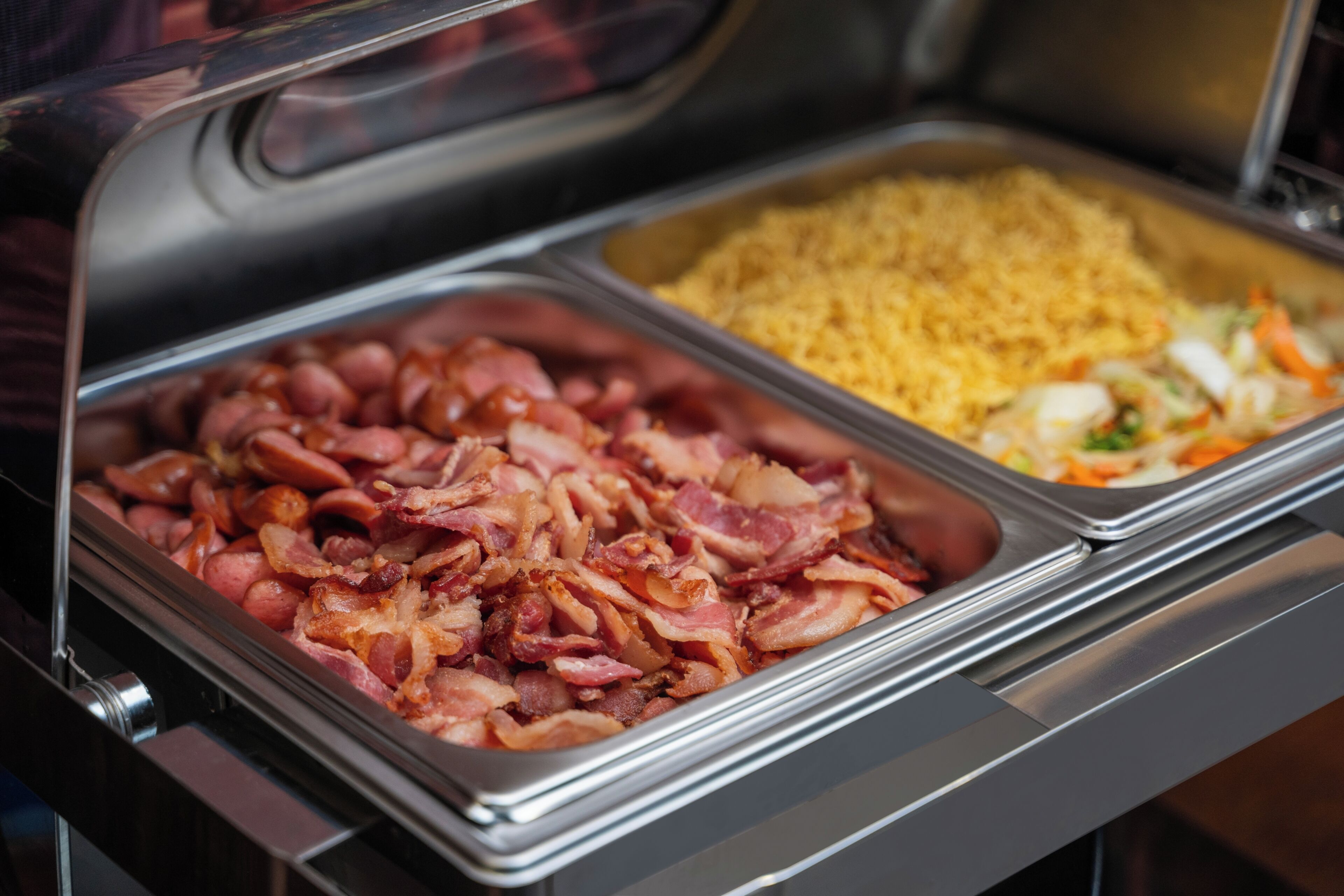 Ein reichhaltiges Buffet mit verschiedenen Speisen, darunter Speck, Reis und einem Gemüsemischung.