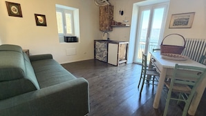 En stue med sofa, spisebord med stole, et tekøkken med vask og køleskab, samt et vindue med udsigt.