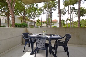 An outdoor patio with a round table and chairs, surrounded by a low wall and trees. - Cozy stay in Lignano (Lignano Sabbiadoro)