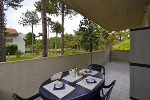 An outdoor dining area with a table set for two, chairs, a small table with a flower vase, and a view of a residential area with trees and a building. - Cozy stay in Lignano (Lignano Sabbiadoro)