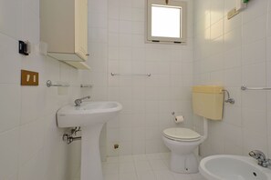 A bathroom with a sink, toilet, and bidet, white tiled walls, and a window. - Adorable apartment with pool (Lignano Sabbiadoro)
