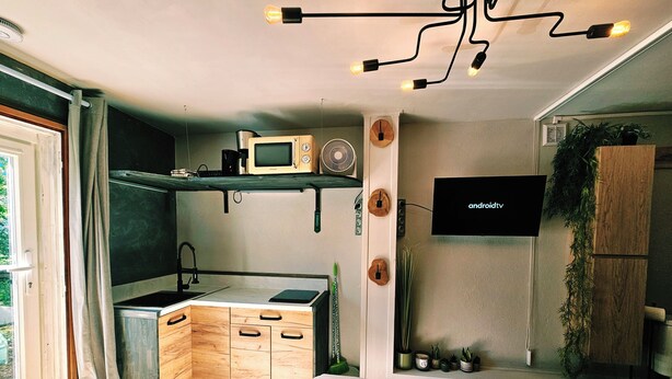 Uma cozinha moderna com micro-ondas, pia e armários de madeira. Há uma televisão montada na parede e um lustre.