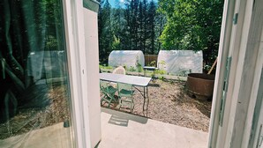 Porta de vidro que dá para uma área externa coberta, com uma mesa e cadeiras, rodeada por árvores e plantas.