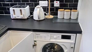 Un espace buanderie compact comprenant un lave-linge, un grille-pain et une bouilloire, le tout posé sur un plan de travail noir.