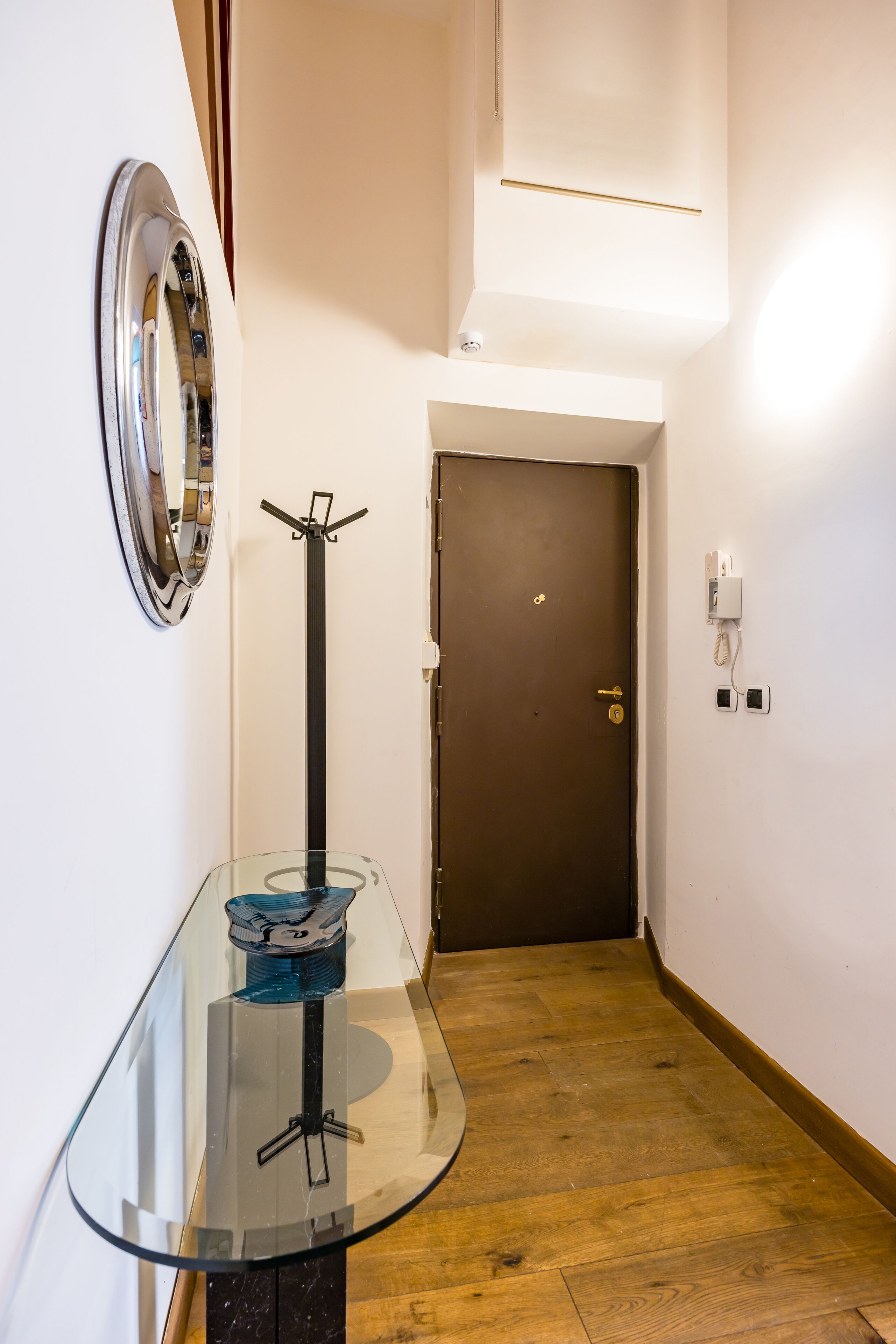 A hallway with a glass table, a coat rack, a door, and a wall-mounted light fixture.