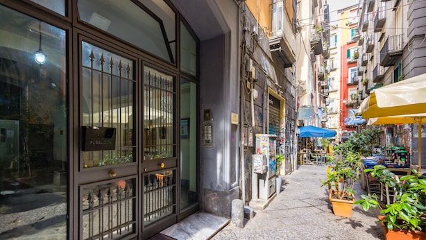 A narrow street with a building featuring large glass doors and a metal gate, potted plants, and outdoor seating under umbrellas. - Quercia Design Home in the Center (Naples)