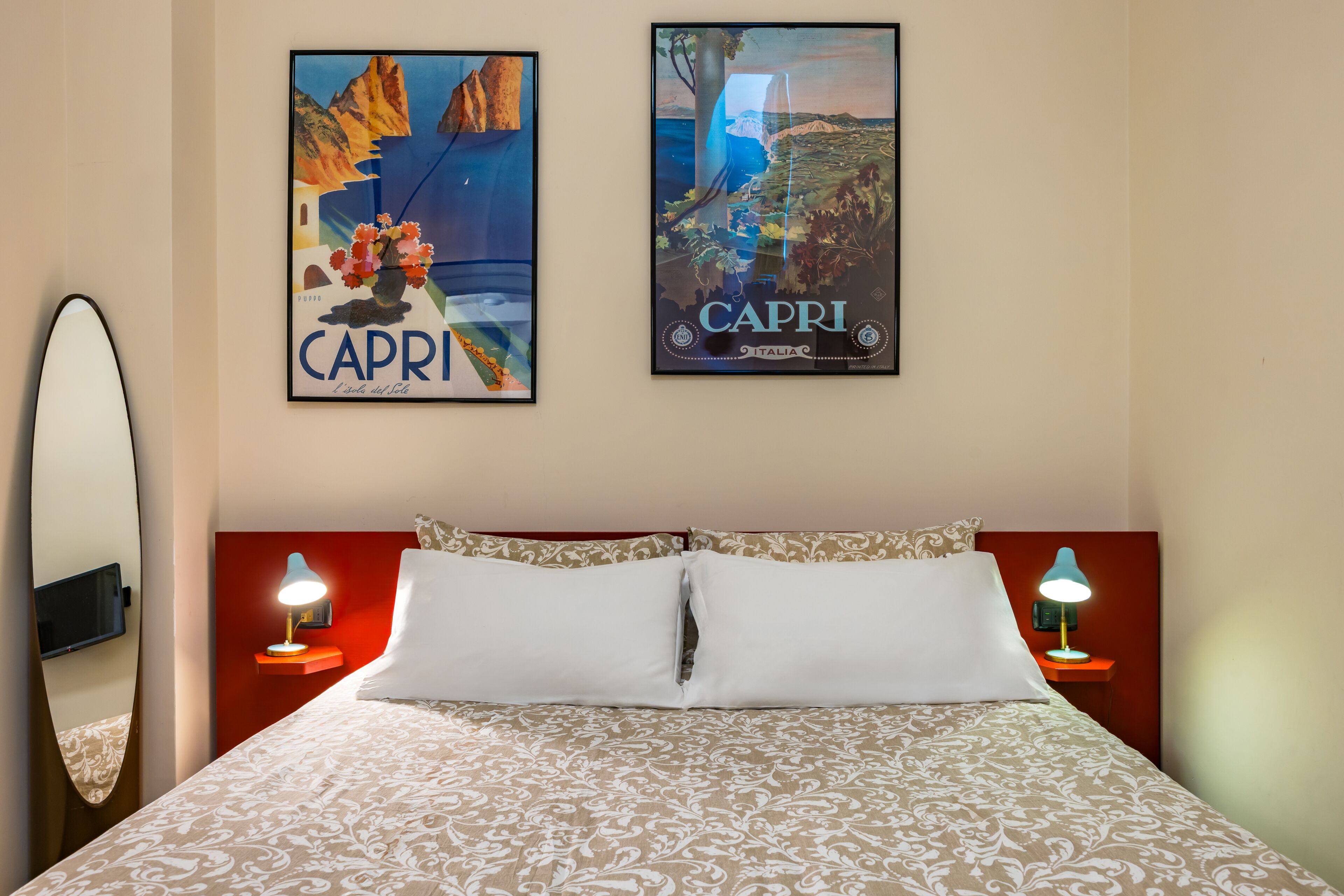A hotel room with a bed, two nightstands with lamps, a large mirror, and two posters on the wall.