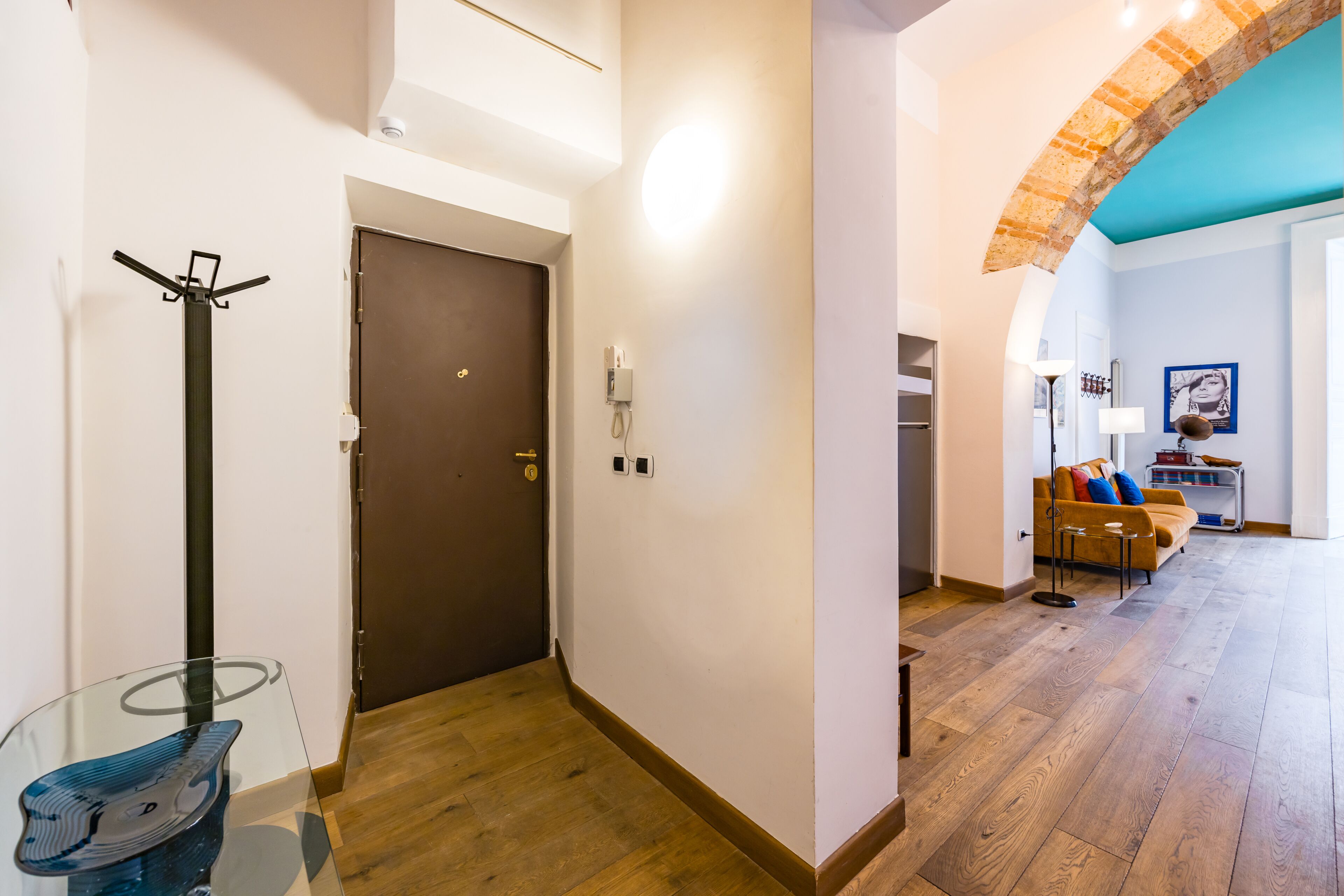 Un pasillo con suelo de madera, una mesa de vidrio, un perchero, una puerta y una sala de estar con sofá y lámpara.