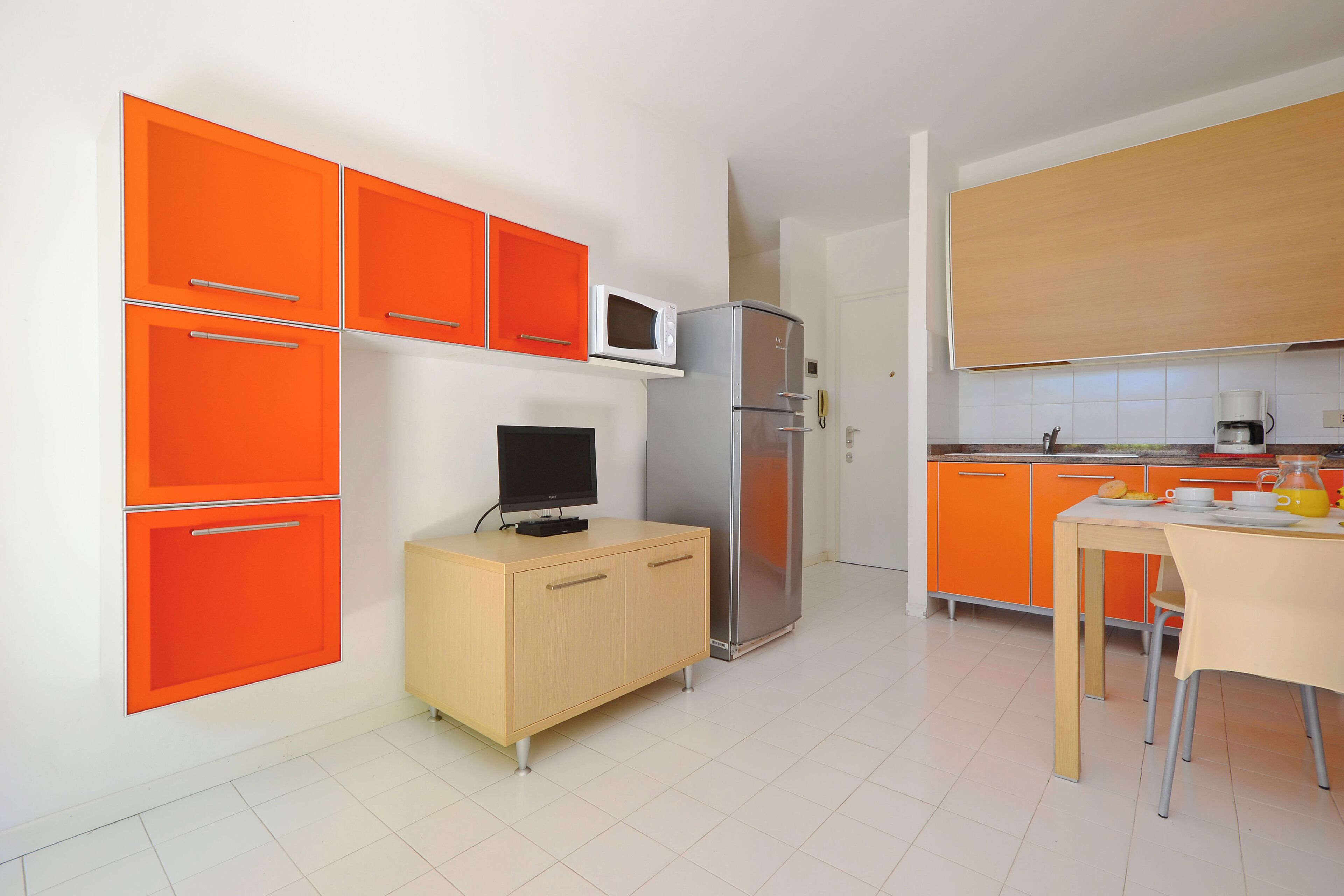 Eine kompakte Küche mit orangefarbenen Schränken, einem Kühlschrank, einer Mikrowelle, einem kleinen Fernseher und einem Essbereich mit Tisch und Stühlen.