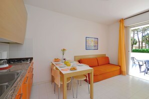 A modern kitchen with a dining table and orange sofa, a painting on the wall, and a view through the glass door. - Bright getaway flat with pool (Lignano Sabbiadoro)