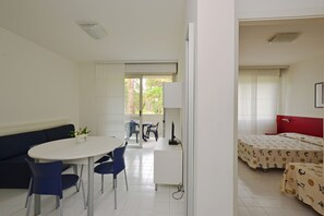 A room with a white desk, blue chairs, a white sofa, and two beds with patterned bedding. - Bright getaway flat with pool (Lignano Sabbiadoro)