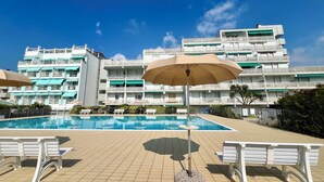 En swimmingpool med liggestole og en parasol, omgivet af en bygning med flere altaner.