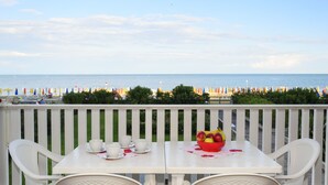 En balkon med et bord dækket til to, med udsigt over en strand med farverige parasoller og krystalklart hav.