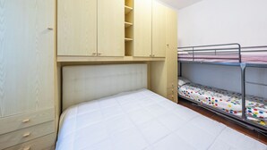 Ein Etagenbett mit weißer Matratze und einem Punktmuster-Bettlaken, ein Holzschrank mit mehreren Fächern und ein Metallbettgestell.