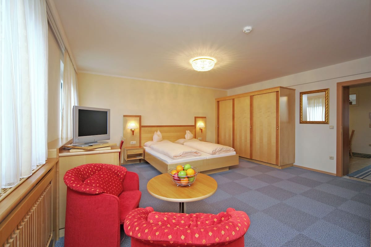 A hotel room with a bed, a television, a small table with fruit, a red chair, and a red ottoman.