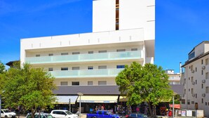 A multi-story building with a modern architectural design, featuring a prominent vertical element and a ground-level shop with a striped awning. - Quiet flat with balcony (Lignano Sabbiadoro)